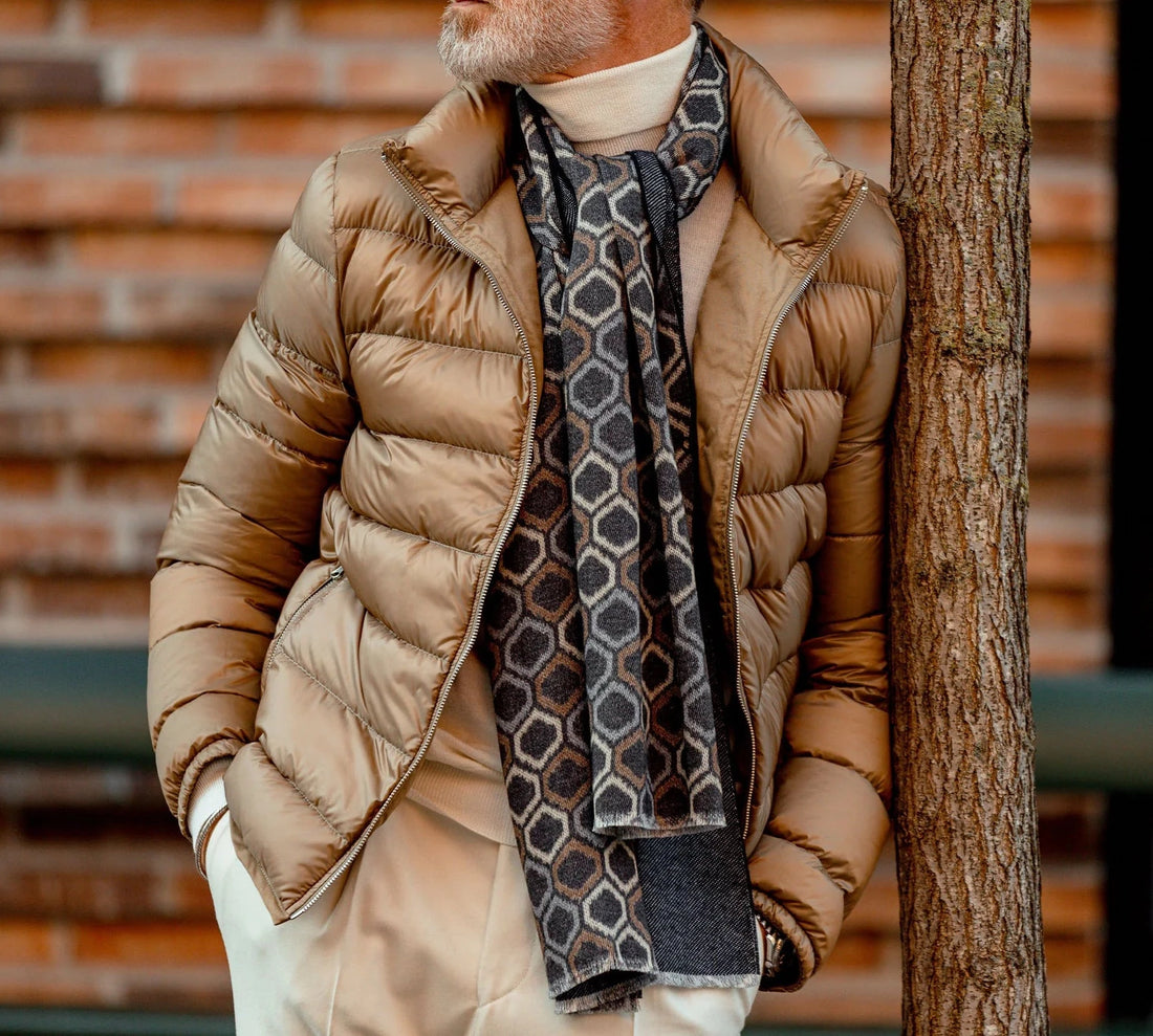 A person wearing a beige quilted jacket and grey turtleneck with a patterned scarf stands by a tree with a brick wall in the background.