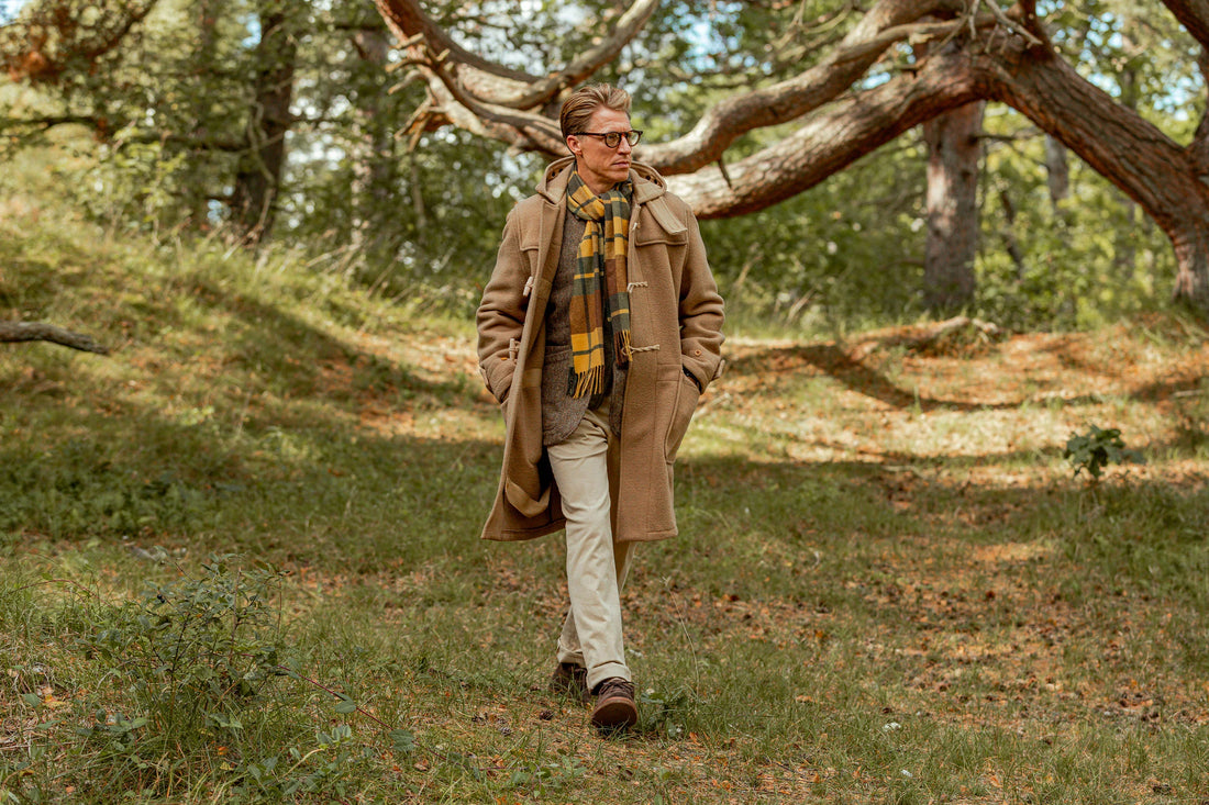 A person in a long coat and scarf walks through a wooded area.