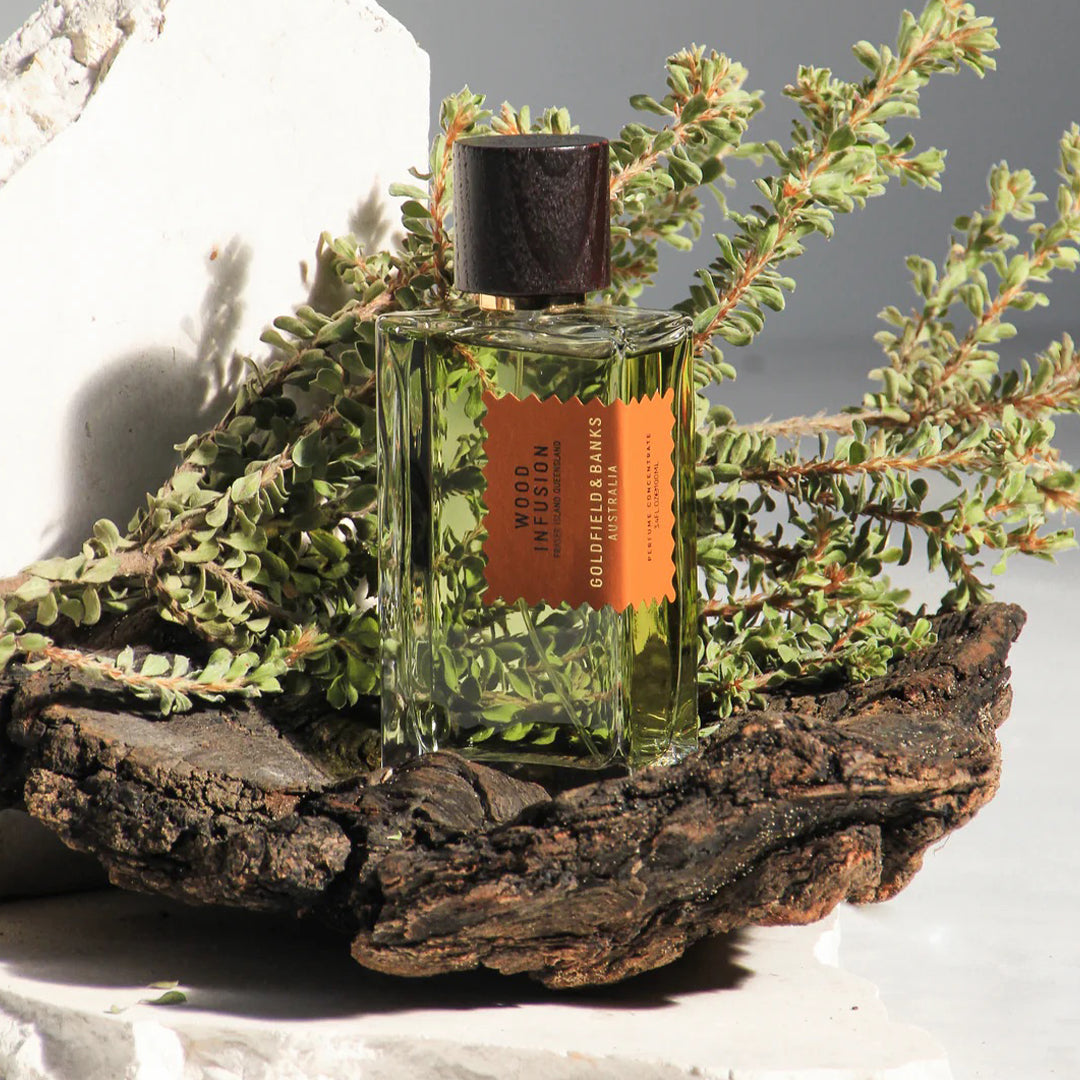 A bottle of Wood Infusion Goldfield & Banks perfume sits on a piece of bark, surrounded by greenery against a neutral background.