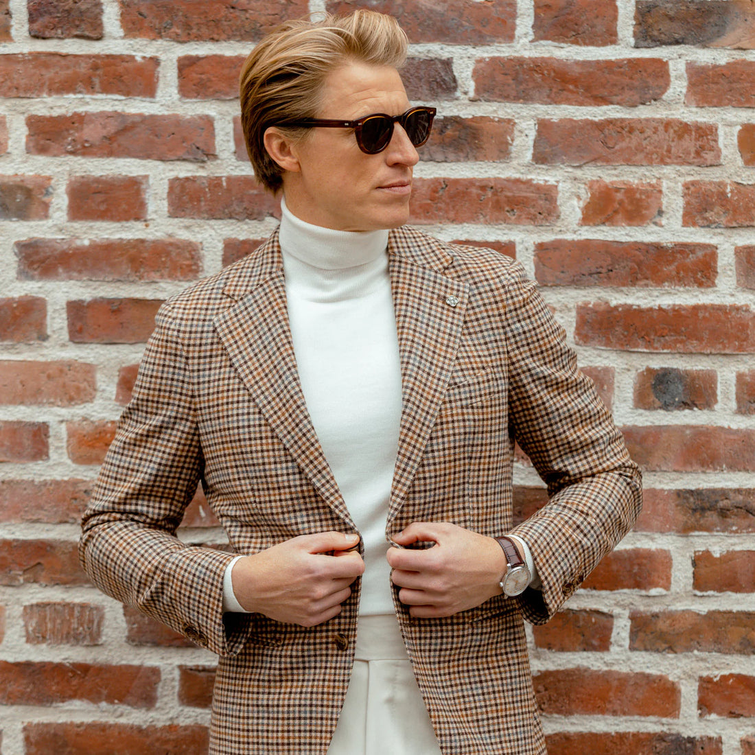 A person in a plaid jacket and turtleneck standing in front of a brick wall.