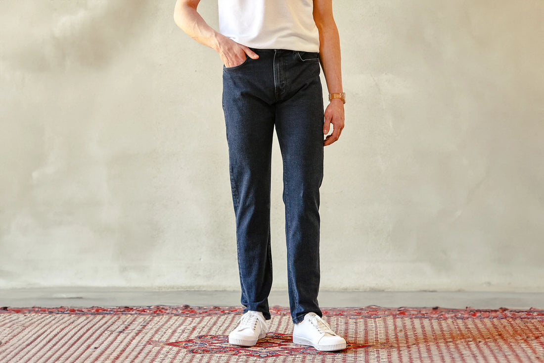 Person standing against a plain background, wearing blue jeans, white sneakers, and a white top.