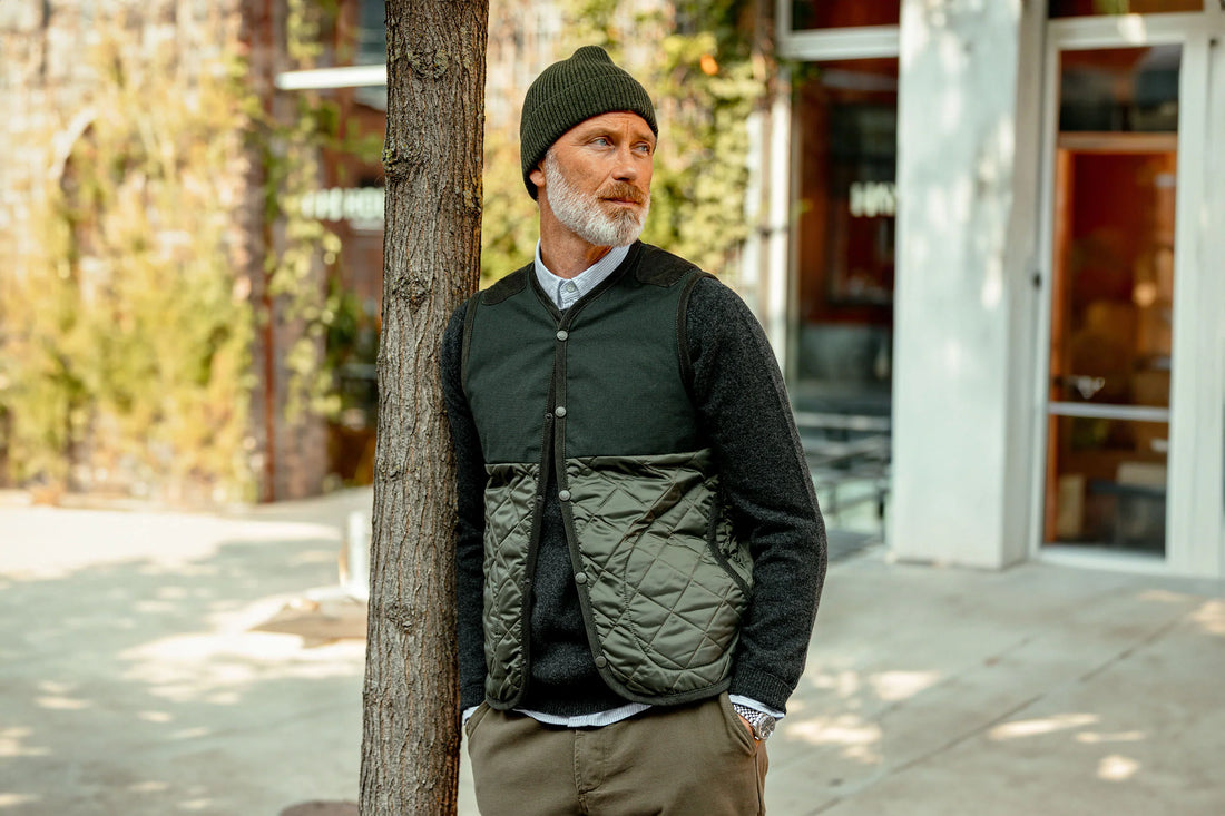A mature man in a beanie and quilted vest leaning against a tree outdoors.