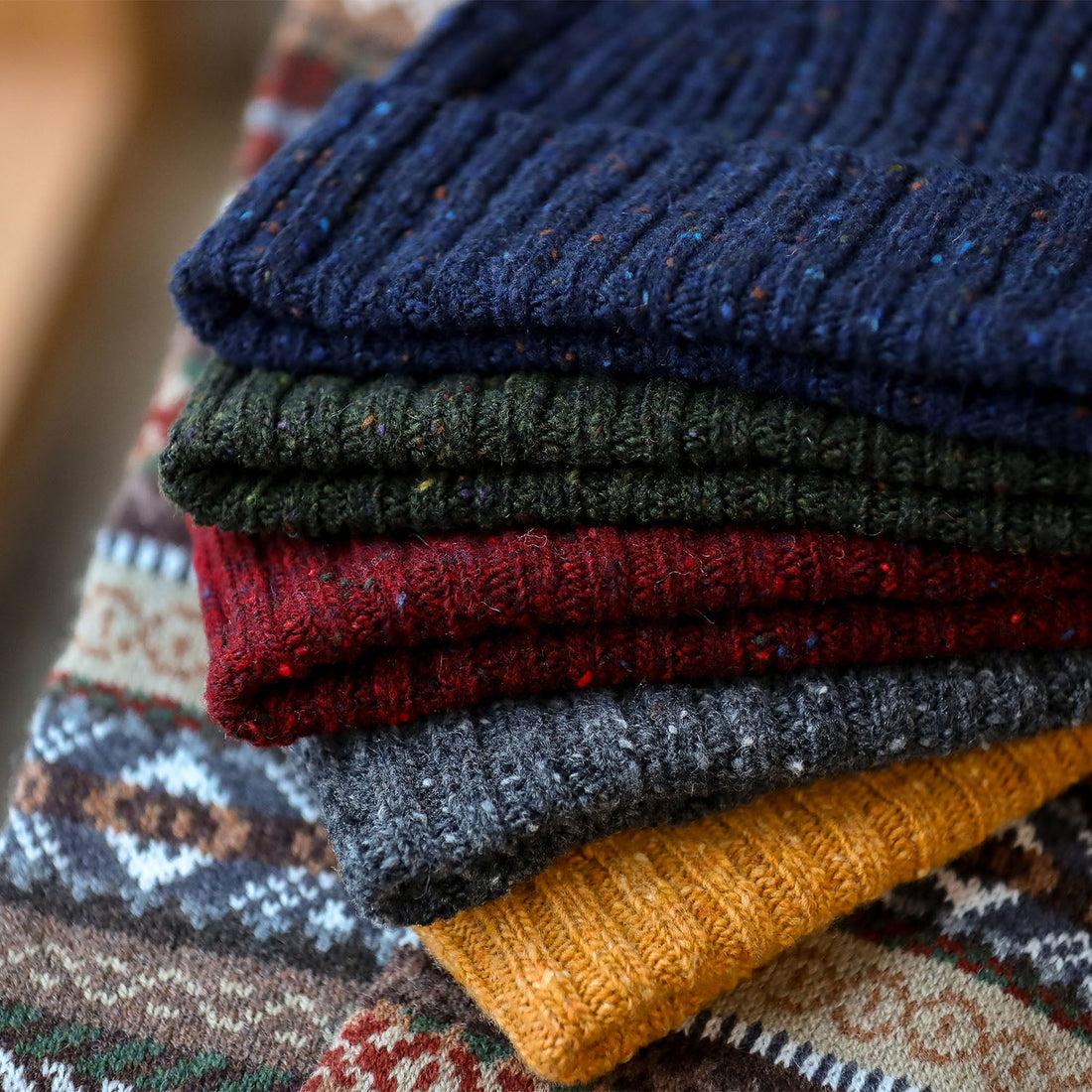 Five folded knit sweaters stacked by color: blue, green, red, gray, and mustard. A patterned textile is visible underneath.