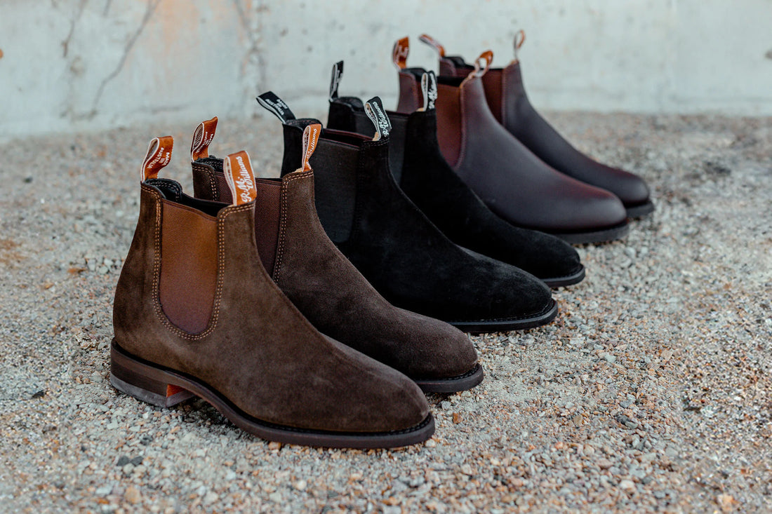 Three pairs of chelsea boots in brown, black, and burgundy lined up on a gravel surface.