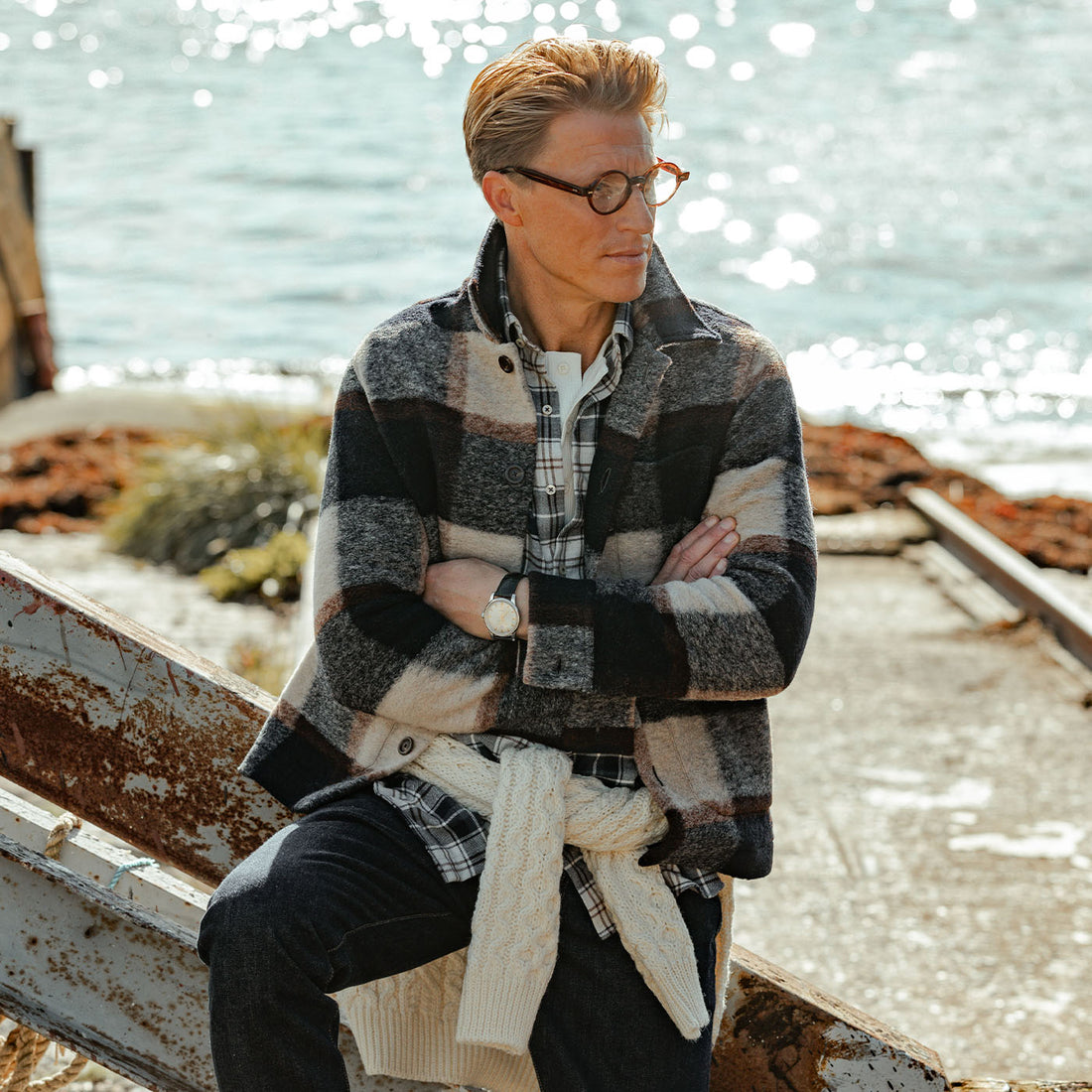 A man in glasses sits on a boat, wearing a plaid jacket and jeans, arms crossed, with sunlight reflecting off the water in the background.