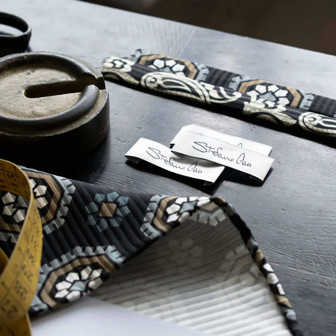 A table with fabric samples, a tape measure, small notes, and a metal object, possibly an ashtray, resting on a dark surface.