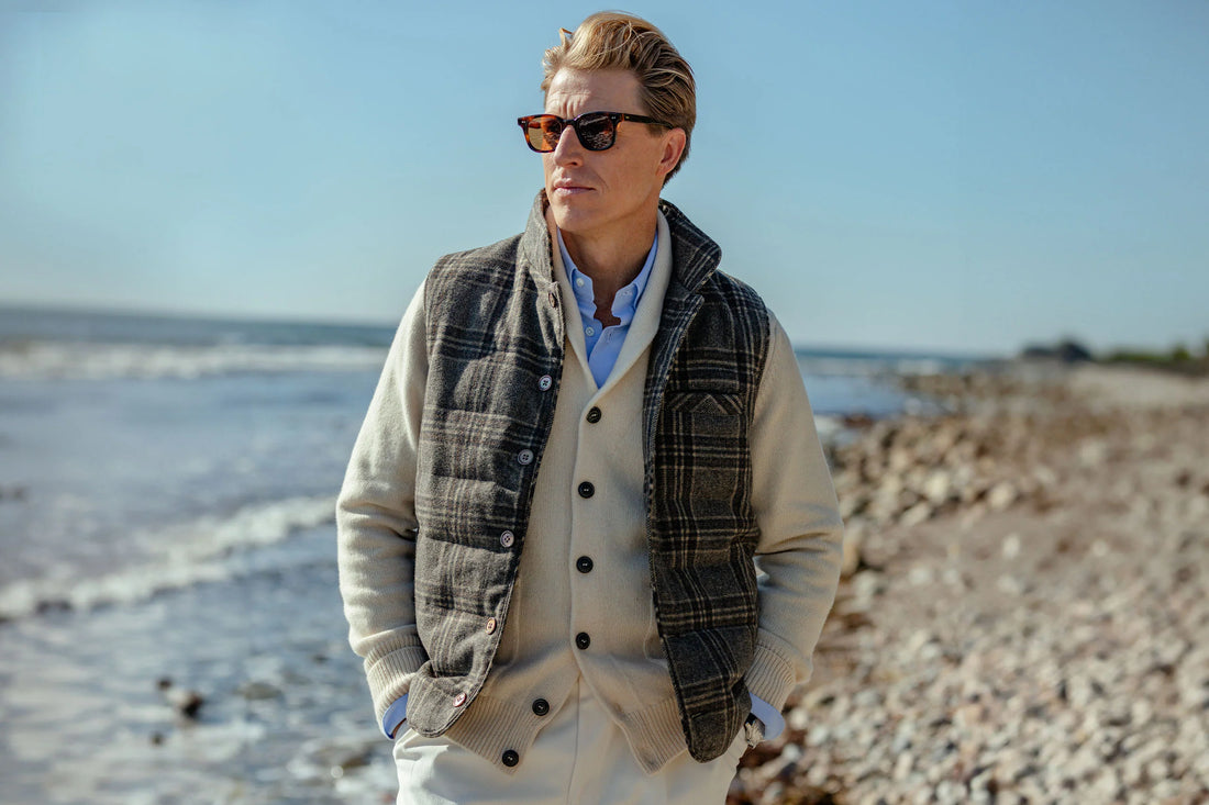 A person standing on a pebble beach near the water, wearing sunglasses and a layered outfit with a checked jacket.
