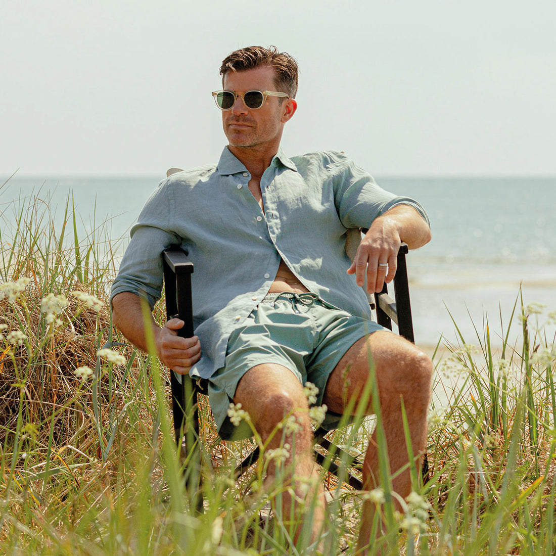 A man in sunglasses sits on a beach chair, wearing a light blue shirt and green shorts, with tall grass and the sea in the background.