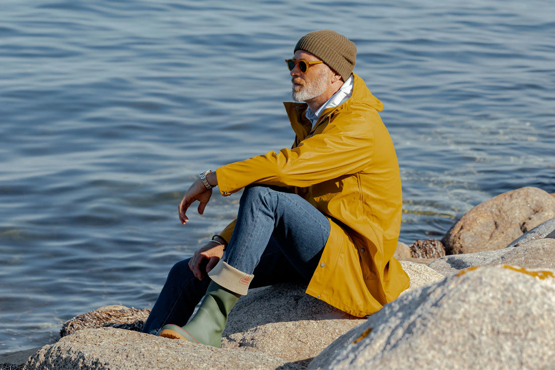 Man in a yellow jacket and beanie sitting on rocks by the water, looking into the distance.
