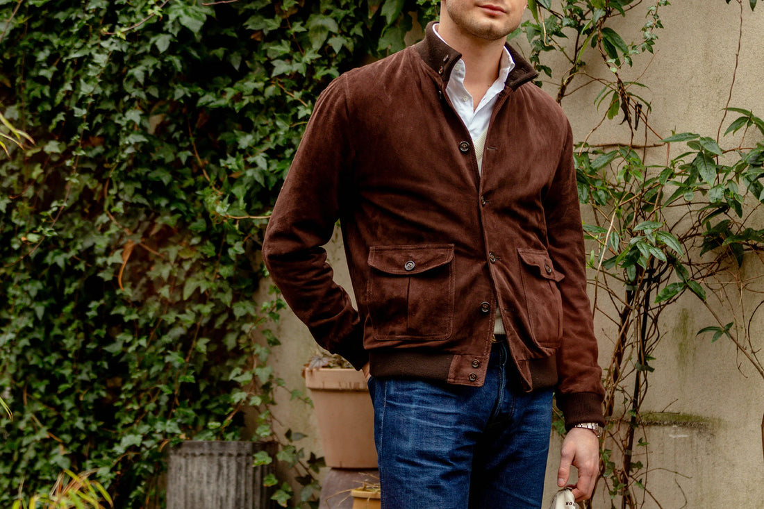 Man in a brown suede jacket and blue jeans standing against a foliage-covered wall.