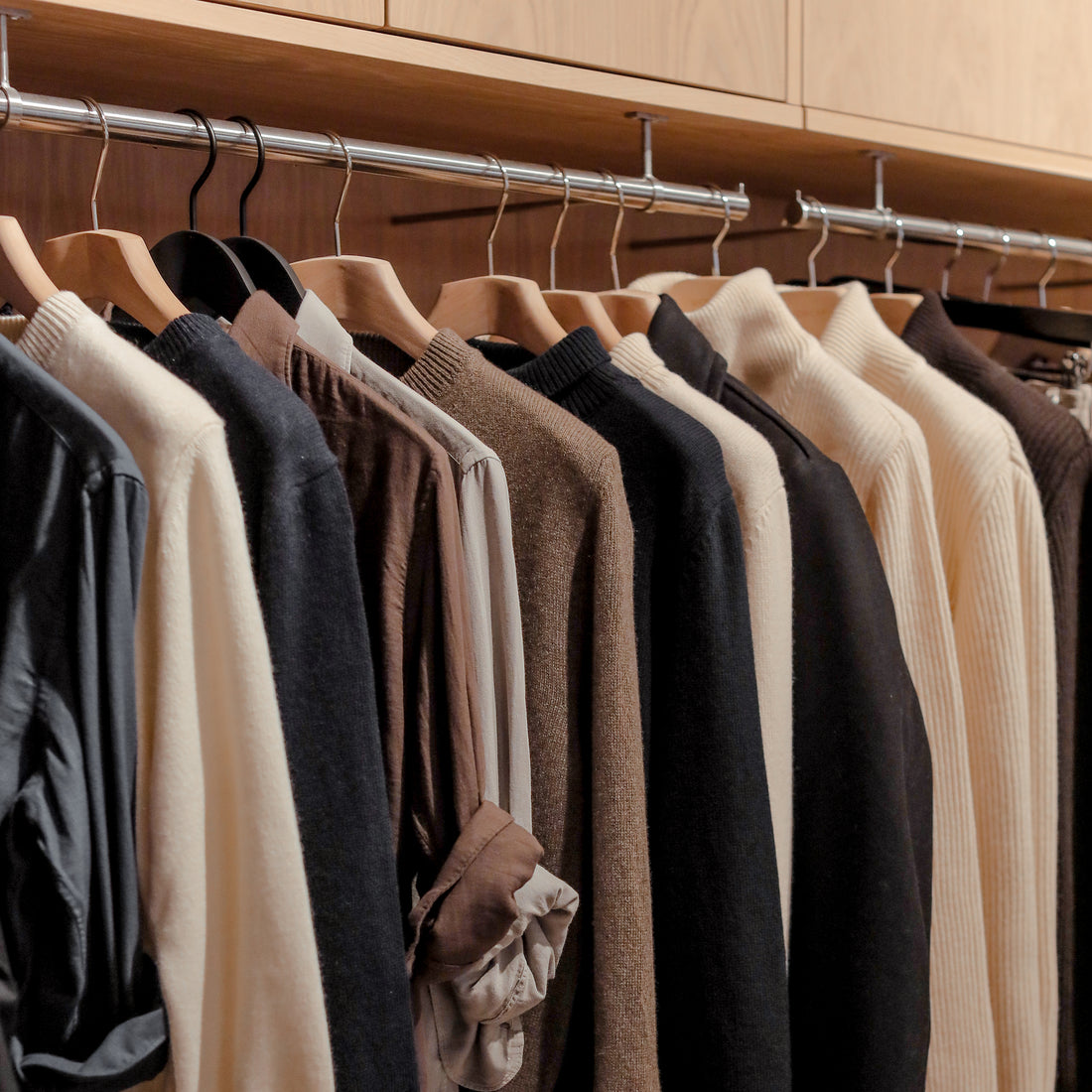 A neatly organized closet with a variety of shirts and sweaters on wooden hangers, arranged by color from light to dark.