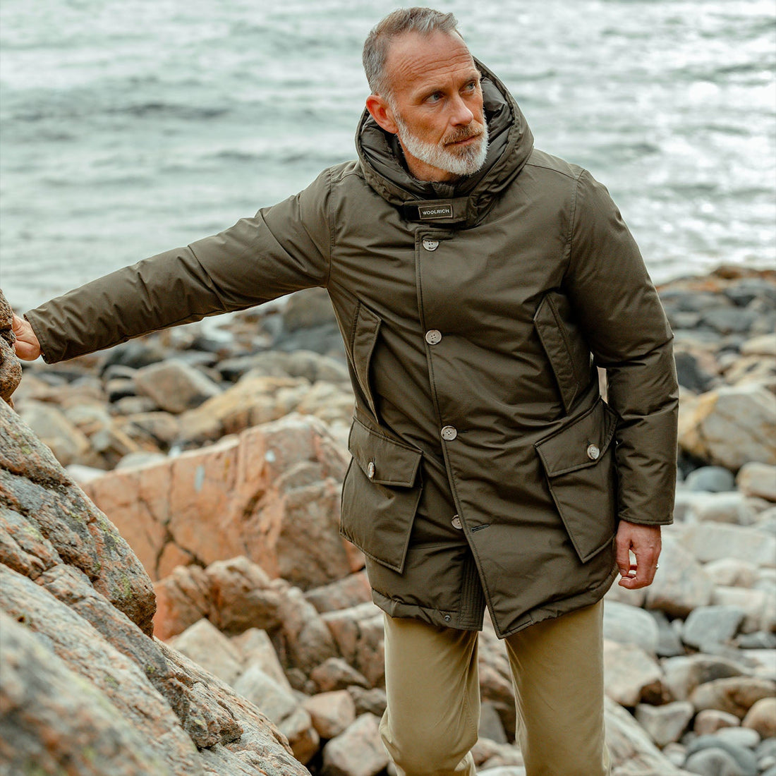 A man in a green parka stands on a rocky shoreline with the ocean in the background.