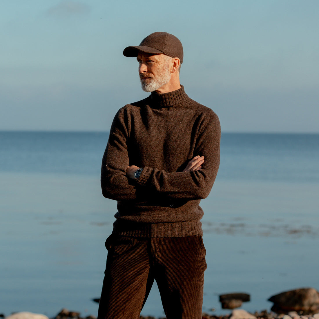 A person in a green sweater and plaid pants stands outdoors with crossed arms, surrounded by a rural landscape.