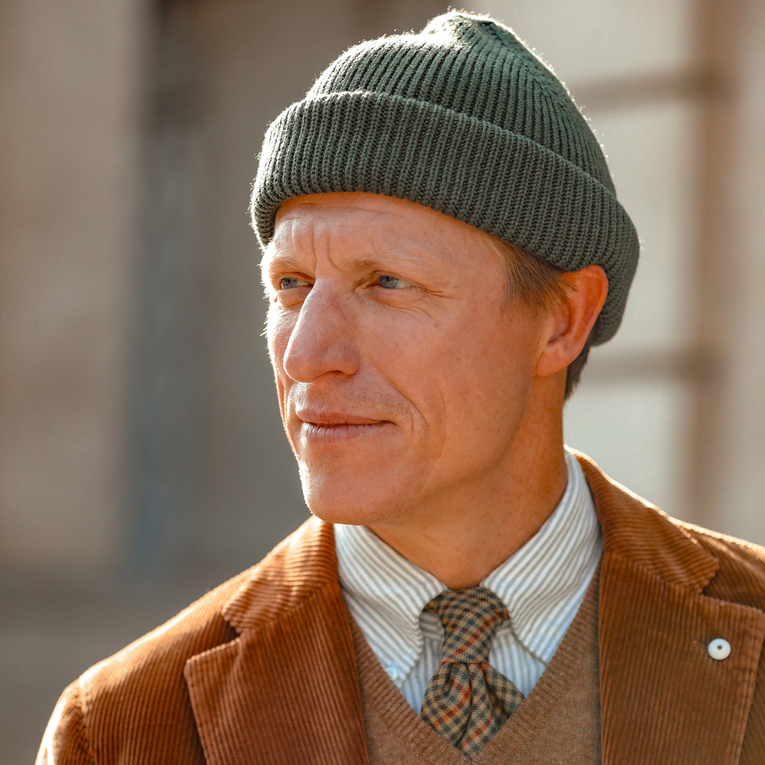 A man wearing a knit beanie, corduroy jacket, striped shirt, and patterned tie looks into the distance.