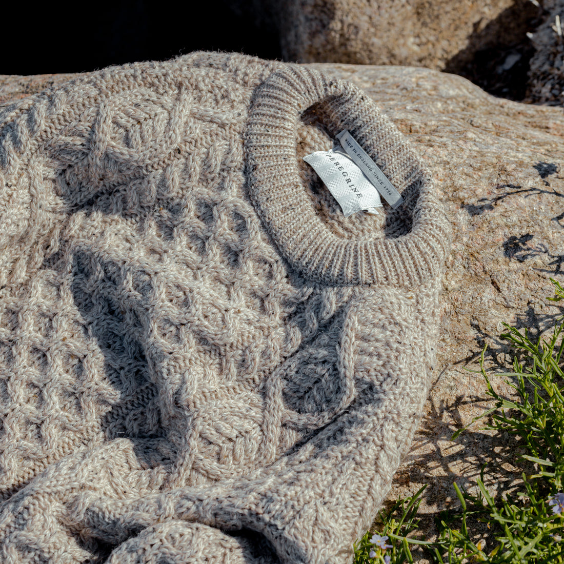 A textured beige sweater with a woven diamond pattern and a visible clothing tag is draped over a rock. Small green plants can be seen in the corner.