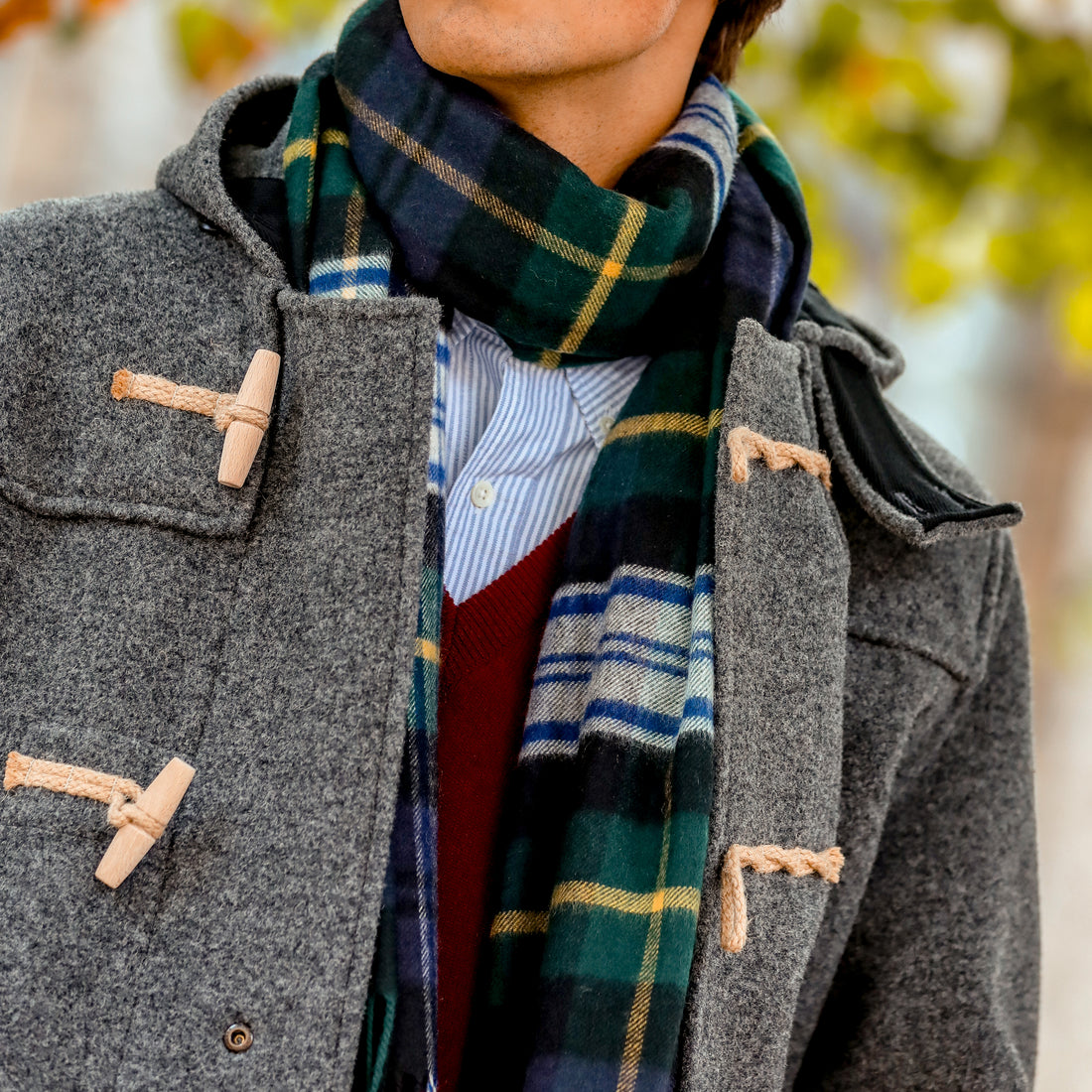 A person is wearing a grey coat with wooden toggle buttons, a striped shirt beneath a red sweater, and a green and blue plaid scarf. The background features blurry greenery.