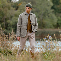 A person in an Olive Melange Harris Tweed Bakers C Jacket by Universal Works and a green beanie walks through a field with greenery and trees in the background.