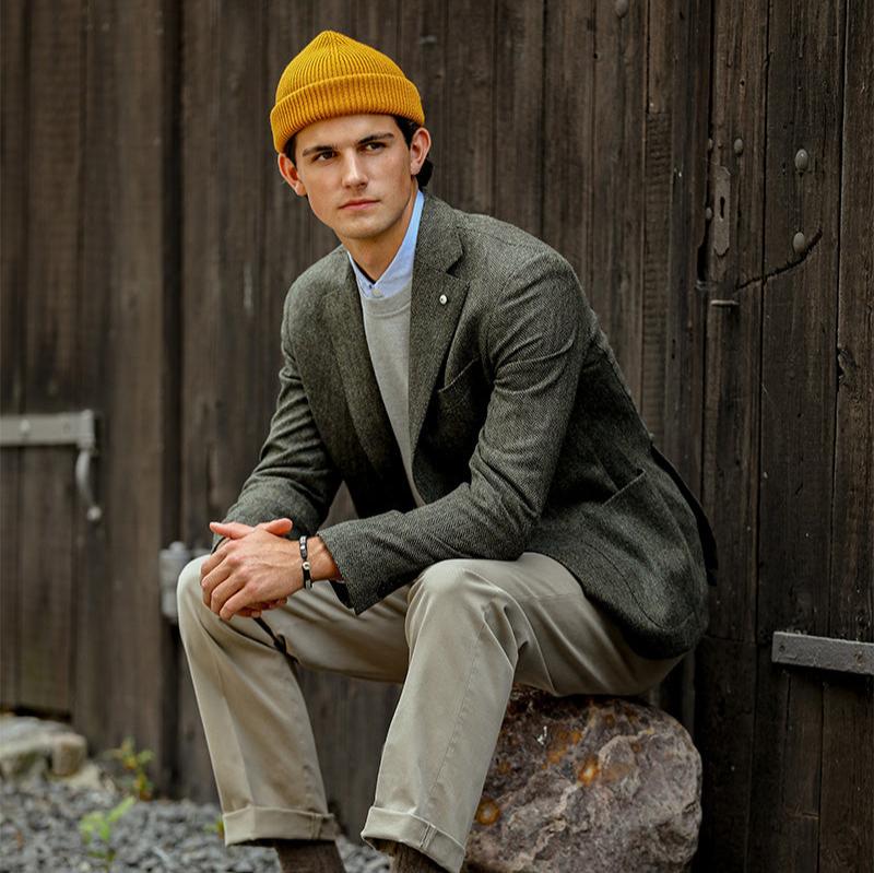 A person leans against a wooden wall, sitting on a rock. They sport a yellow beanie and beige pants, complemented by brown shoes and a Dark Green Wool Cashmere Herringbone Blazer from L.B.M. 1911, reflecting Italian tailoring craftsmanship.