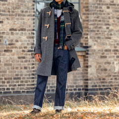 A person stands outdoors in front of a brick wall, dressed in a gray duffle coat and blue jeans. Their jeans, the Dark Blue Cotton 714 One Wash Jeans by Resolute, are crafted from unsanforized selvedge denim and feature a tapered slim leg for a contemporary fit. They complete the look with a scarf and brown shoes.