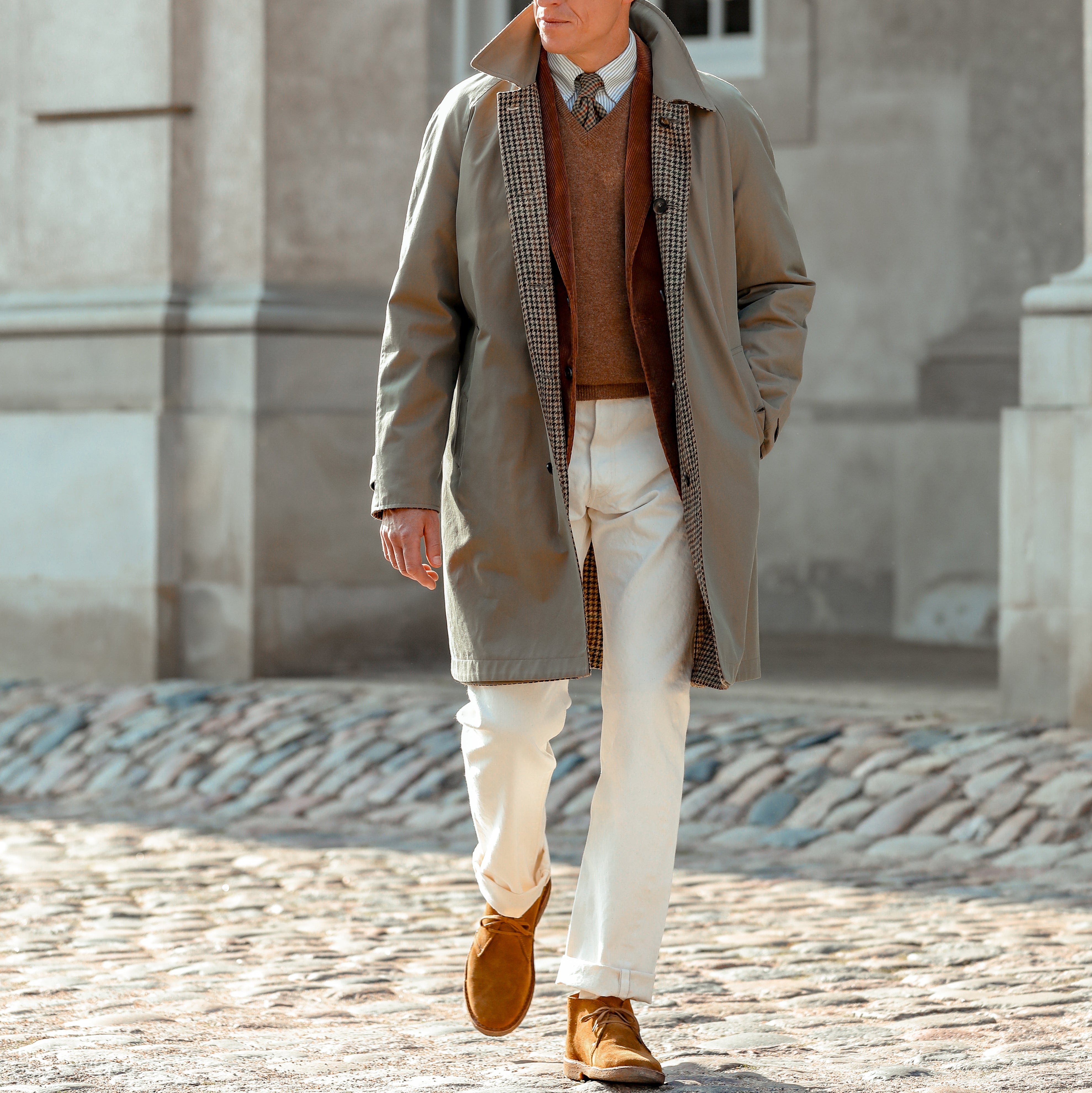 A person wearing a long tan coat, brown sweater, Resolute's Cream Cotton Selvedge 714 One Wash Jeans, and brown shoes walks outdoors on a cobblestone path, with an architectural structure in the background.