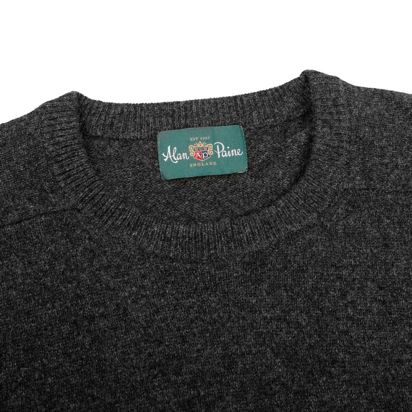 A close-up of the Charcoal Grey Lambswool Crew Neck sweater from Alan Paine, featuring a green label that reads "Alan Paine, England," made from the finest Australian lambswool.