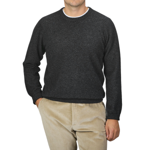 A man is wearing an Alan Paine Charcoal Grey Lambswool Crew Neck sweater along with light brown corduroy pants, standing against a plain gray background with his right hand in his pocket.