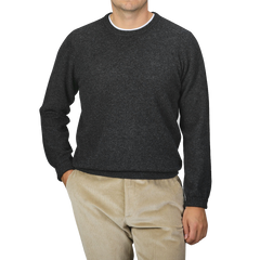 A man is wearing an Alan Paine Charcoal Grey Lambswool Crew Neck sweater along with light brown corduroy pants, standing against a plain gray background with his right hand in his pocket.