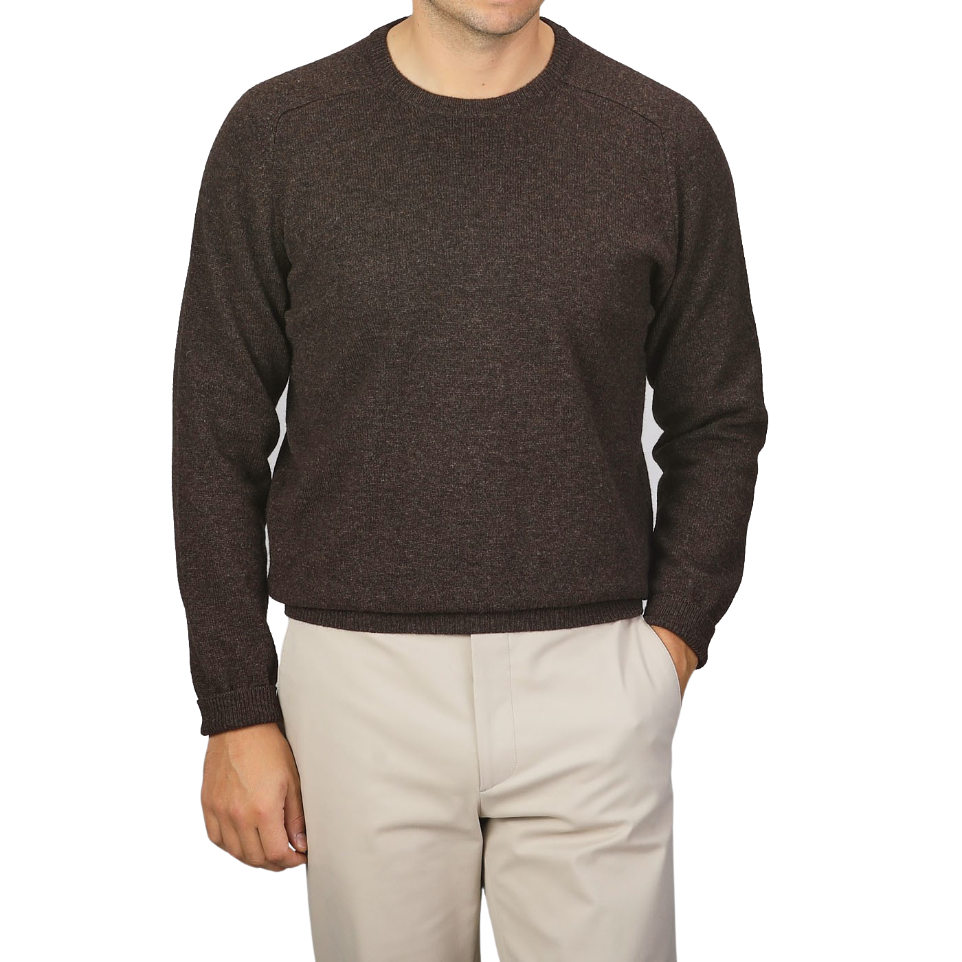 A person is wearing an Alan Paine Cocoa Brown Lambswool Crew Neck sweater and beige pants. The person's left hand is in their pocket. The background is plain light gray.