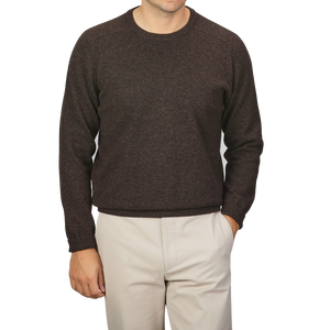 A person is wearing an Alan Paine Cocoa Brown Lambswool Crew Neck sweater and beige pants. The person's left hand is in their pocket. The background is plain light gray.