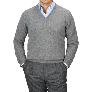 A person wearing an Alan Paine Grey Melange Lambswool V-Neck sweater over a light blue collared shirt, with hands in the pockets of grey pants. The background is plain and grey.