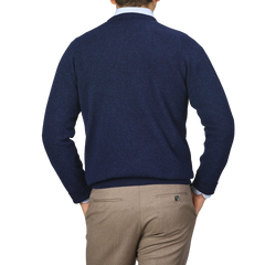 An individual is wearing an Alan Paine Indigo Blue Lambswool V-Neck sweater and beige pants, standing with his back to the camera, hands in pockets, against a plain gray background.