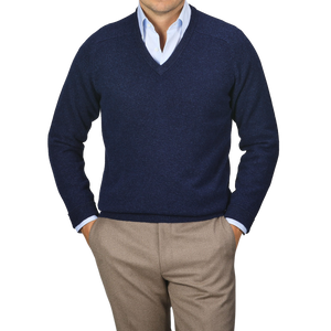 A person wearing an Alan Paine Indigo Blue Lambswool V-Neck sweater over a light blue dress shirt and beige pants, standing with hands in pockets against a plain background.