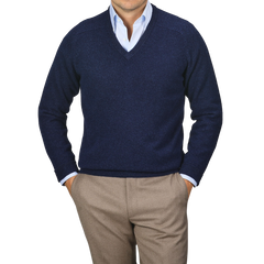 A person wearing an Alan Paine Indigo Blue Lambswool V-Neck sweater over a light blue dress shirt and beige pants, standing with hands in pockets against a plain background.