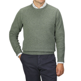 A man wearing an Alan Paine Landscape Green Lambswool Crew Neck sweater over a white collared shirt and dark blue pants stands against a plain gray background.