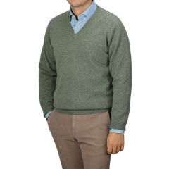 A person is wearing an Alan Paine Landscape Green Lambswool V-Neck sweater over a light blue collared shirt, paired with beige pants. The individual's face is not shown in the image.