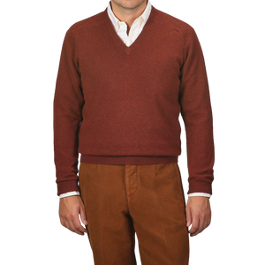 A person wearing an Alan Paine Nebula Red Brown Lambswool V-Neck sweater over a white button-down shirt, paired with brown pants. The background is plain gray.