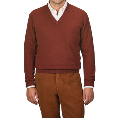 A person wearing an Alan Paine Nebula Red Brown Lambswool V-Neck sweater over a white button-down shirt, paired with brown pants. The background is plain gray.