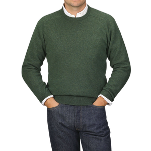 A person wearing an Alan Paine Rosemary Green Lambswool Crew Neck over a white shirt with blue jeans, standing with their hands in their pockets against a plain background.