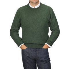 A person wearing an Alan Paine Rosemary Green Lambswool Crew Neck over a white shirt with blue jeans, standing with their hands in their pockets against a plain background.