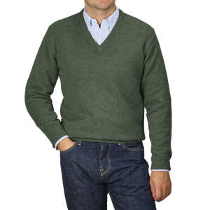 A person wearing an Alan Paine Rosemary Green Lambswool V-Neck, paired with a blue striped shirt and blue jeans, stands against a neutral background.