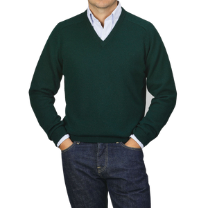 A person dressed in an Alan Paine Tartan Green Lambswool V-Neck sweater layered over a light blue collared shirt, paired with dark jeans, stands with their hands in their pockets against a plain gray background.