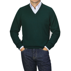 A person dressed in an Alan Paine Tartan Green Lambswool V-Neck sweater layered over a light blue collared shirt, paired with dark jeans, stands with their hands in their pockets against a plain gray background.