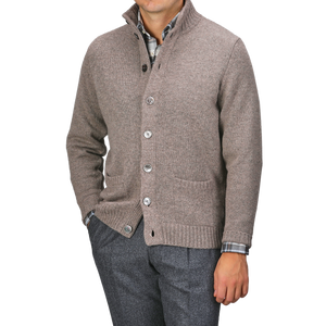 A person wearing an Alan Paine Taupe Beige Lambswool Landford Cardigan over a plaid shirt and gray trousers, with hands in pockets.