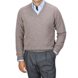 A person wearing an Alan Paine Vole Beige Lambswool V-Neck sweater over a blue dress shirt with updated fit gray trousers stands against a plain background.