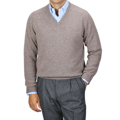 A person wearing an Alan Paine Vole Beige Lambswool V-Neck sweater over a blue dress shirt with updated fit gray trousers stands against a plain background.