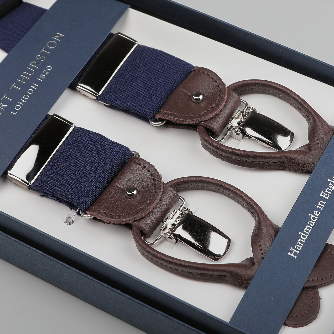 Box containing blue wool fresco braces and brown leather suspenders with silver clasps, branded as the esteemed British heritage brand "Albert Thurston" and labeled "Handmade in England.