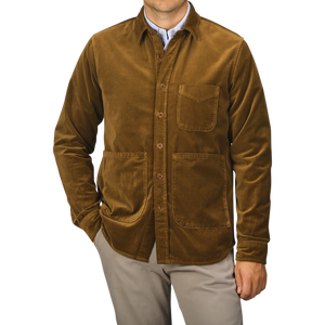 A person wearing the Aspesi Tobacco Brown Cotton Velvet Overshirt, featuring three front pockets and a collared design, stands with hands in pockets against a plain background. This ensemble, expertly crafted from pure cotton velvet, showcases the refined touch of an Italian outerwear specialist.