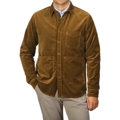 A person wearing the Aspesi Tobacco Brown Cotton Velvet Overshirt, featuring three front pockets and a collared design, stands with hands in pockets against a plain background. This ensemble, expertly crafted from pure cotton velvet, showcases the refined touch of an Italian outerwear specialist.