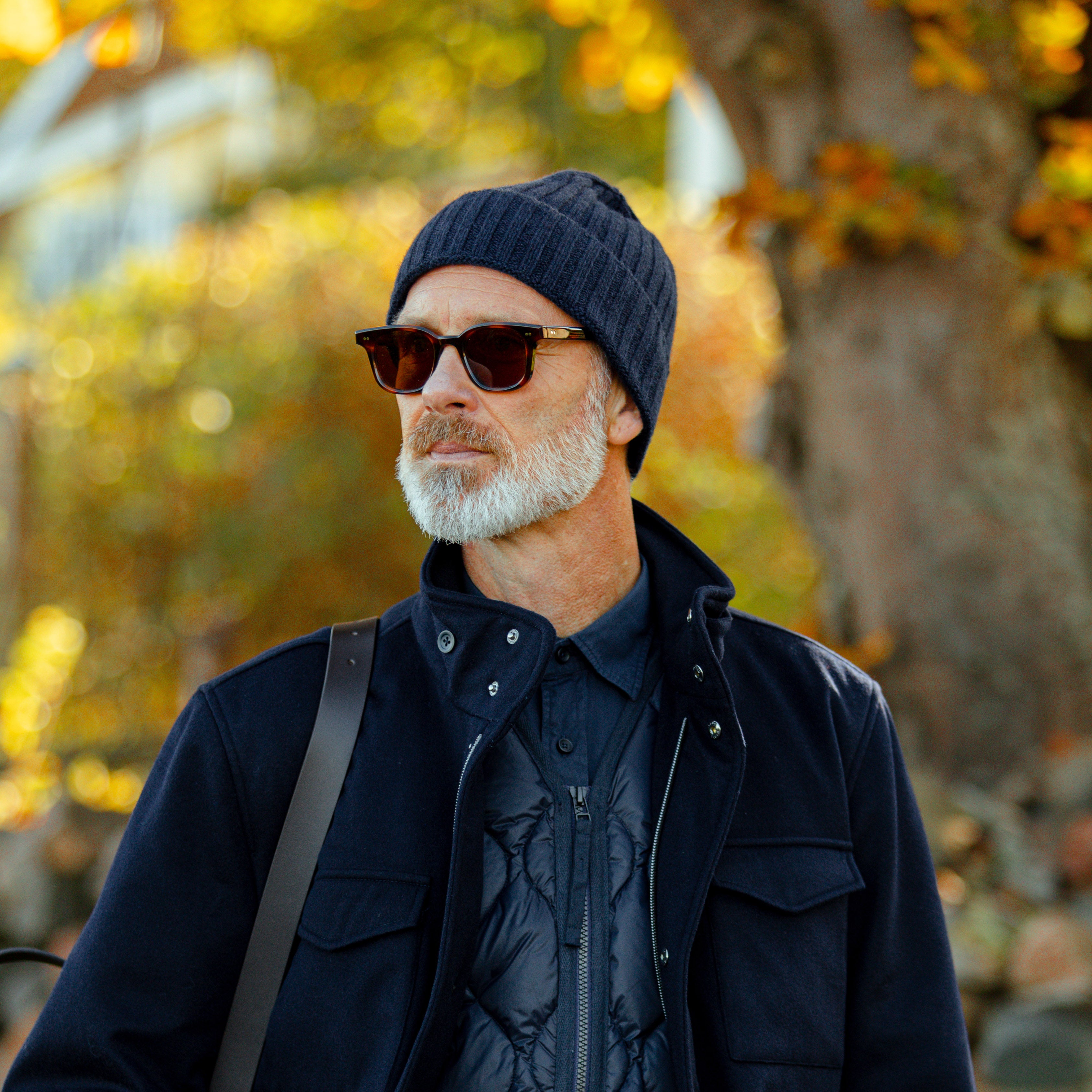 A man with a beard in a dark jacket, wearing a Navy Blue Ribbed Cashmere Beanie by Daniele Fiesoli and sunglasses, stands outdoors in a tree-lined area with autumn leaves.