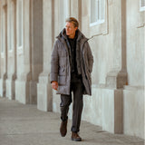 A person wearing a long, taupe beige Montecore Nylon Down Padded Parka and dark pants is walking outside next to a building with large columns.